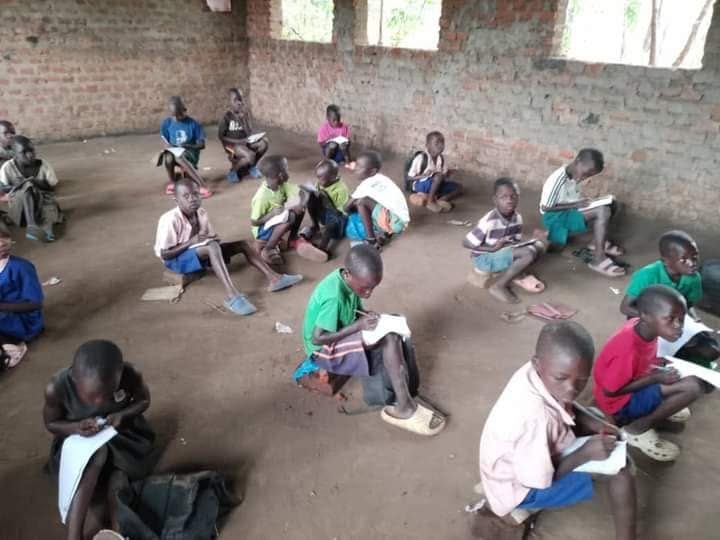 Children sit on the floor of a bare classroom, writing in exercise books on their laps