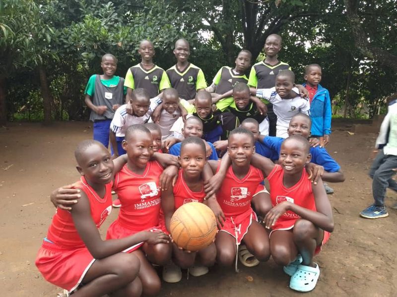 A girls' team in red kit kneels in front with a ball, with more children standing behind them