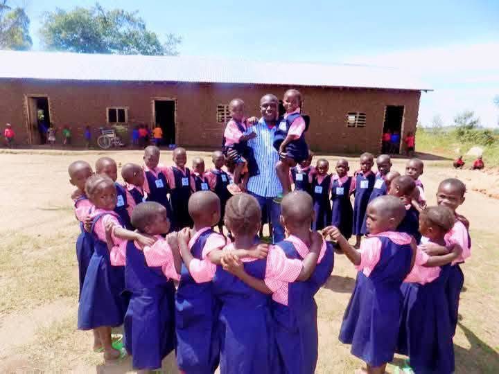 Young pupils in blue and pink uniforms stand in a circle with their teacher in front of a brick classroom block